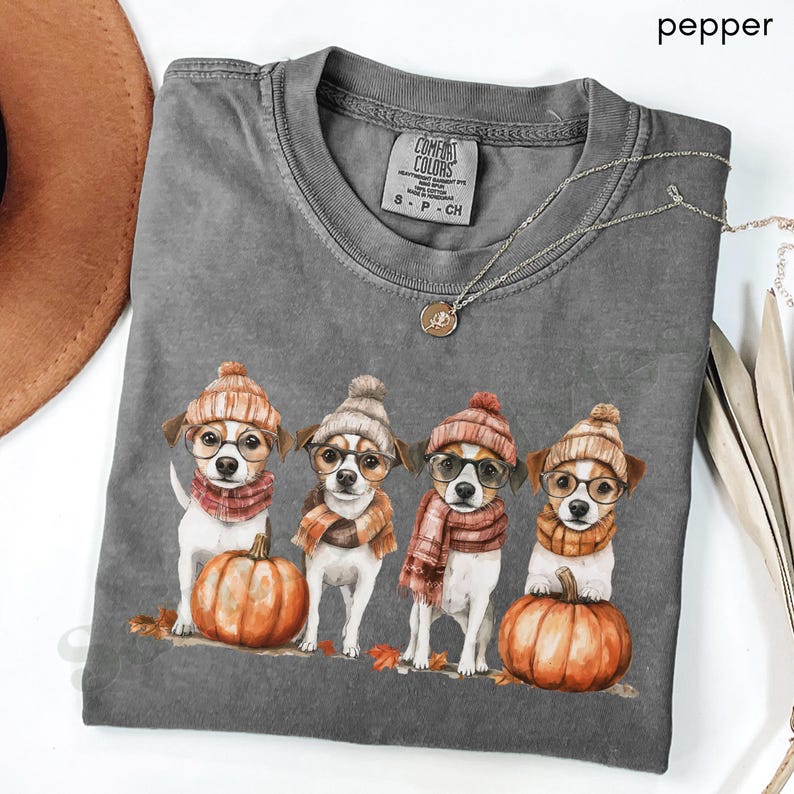 K&ouml;nnte beinhalten: Ein graues Comfort Colors T-Shirt mit einer Grafik von vier Hunden, die H&uuml;te, Brillen und Schals tragen und neben K&uuml;rbissen stehen. Das Wort "pepper" befindet sich in der oberen rechten Ecke. Eine goldene Halskette ist &uuml;ber dem Shirt drapiert.