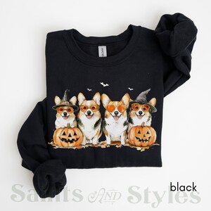 May include: Black sweatshirt with a Halloween-themed design featuring four corgis wearing witch hats and sunglasses, holding pumpkins. The corgis are arranged in a row, with the pumpkins in front of them.
