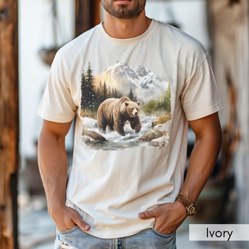 Puede incluir: Una camiseta beige con un estampado gr&aacute;fico de un oso marr&oacute;n caminando por un paisaje monta&ntilde;oso nevado.