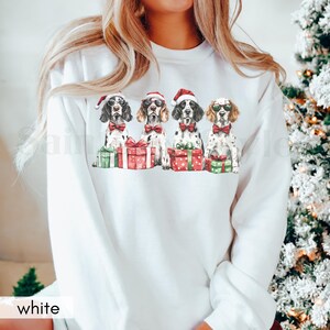 May include: White sweatshirt with a Christmas-themed design featuring four dogs wearing Santa hats and sunglasses, holding presents. The dogs are all wearing red and green Christmas colors.