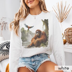 Op de afbeelding: Witte trui met een grafische print van een orang-oetan die in een jungle-omgeving zit met een blauw-rode papegaai die erboven vliegt.