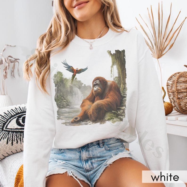 Op de afbeelding: Witte trui met een grafische print van een orang-oetan die in een jungle-omgeving zit met een blauw-rode papegaai die erboven vliegt.