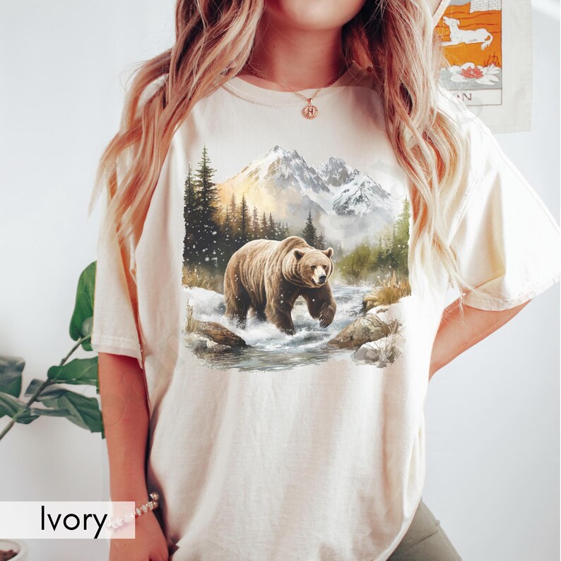 Puede incluir: Una camiseta beige con un estampado gr&aacute;fico de un oso marr&oacute;n caminando por un paisaje monta&ntilde;oso nevado. El oso camina por un arroyo con monta&ntilde;as nevadas en el fondo.