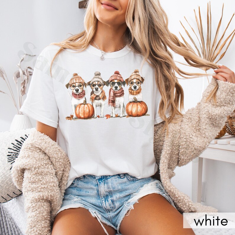 K&ouml;nnte beinhalten: Wei&szlig;es T-Shirt mit einer Aquarellillustration von vier Hunden, die H&uuml;te, Schals und Brillen tragen und auf K&uuml;rbissen sitzen. Die Hunde sind von Herbstbl&auml;ttern umgeben. Das Wort "white" befindet sich in der unteren rechten Ecke.