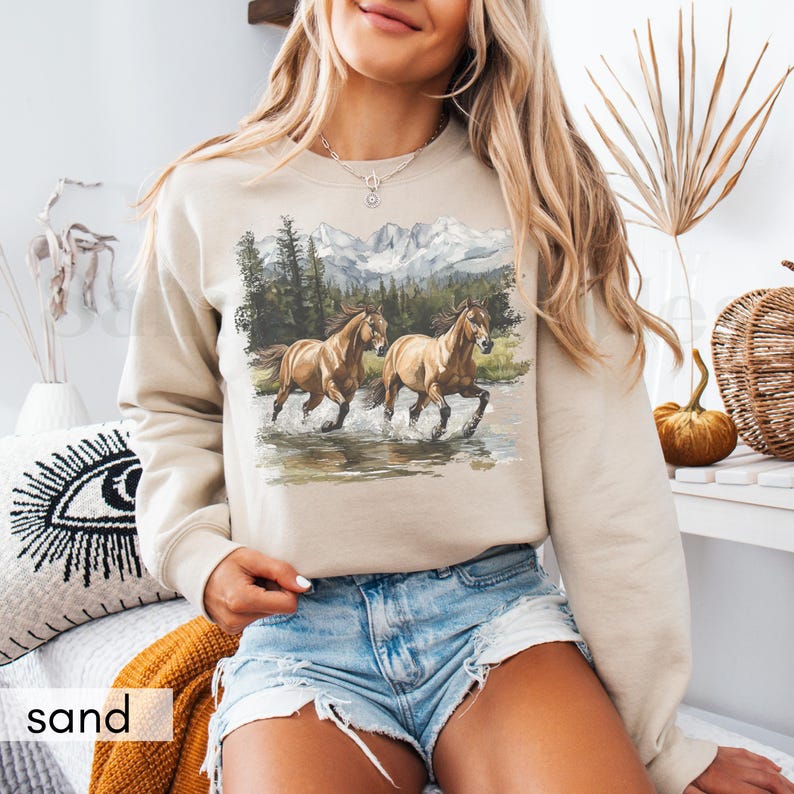 May include: A beige sweatshirt with a graphic print of two brown horses running through a mountain landscape. The horses are in a stream of water and the mountains are in the background.