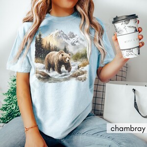 Puede incluir: Una camiseta azul claro con un gr&aacute;fico de un oso marr&oacute;n caminando por un arroyo de monta&ntilde;a nevado. Las monta&ntilde;as est&aacute;n en el fondo y el oso est&aacute; en primer plano. La camiseta est&aacute; hecha de una tela suave y c&oacute;moda.