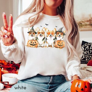 May include: White sweatshirt with a graphic of four corgis wearing witch hats and sunglasses, holding pumpkins. The corgis are in a row, facing forward. The sweatshirt has a crew neck and long sleeves.
