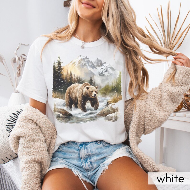 Puede incluir: Camiseta blanca con un gr&aacute;fico de un oso marr&oacute;n caminando por un paisaje monta&ntilde;oso nevado.