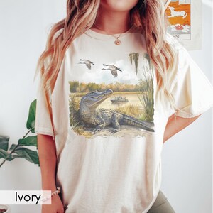 May include: Ivory-colored t-shirt featuring a detailed illustration of an alligator resting near a body of water. Two birds fly above the alligator, with a boat in the background. The shirt has a relaxed fit.