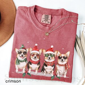 May include: A crimson red t-shirt featuring a graphic of four corgis wearing Santa hats and sunglasses. The corgis are sporting scarves and are standing in front of a holly sprig. The shirt has the text "COMFORT COLORS" on the tag.