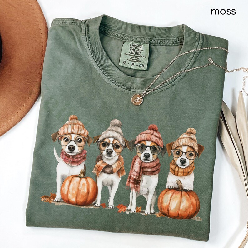 K&ouml;nnte beinhalten: Moosgr&uuml;nes Comfort Colors T-Shirt mit einer Grafik von vier Hunden, die H&uuml;te, Brillen und Schals tragen, jeder neben einem orangefarbenen K&uuml;rbis. Das Shirt hat einen Rundhalsausschnitt und kurze &Auml;rmel. Eine goldene Halskette ist &uuml;ber dem Shirt drapiert.