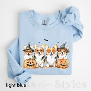 May include: Light blue sweatshirt with a graphic of four corgis wearing witch hats and sunglasses, holding pumpkins with jack-o'-lantern faces.