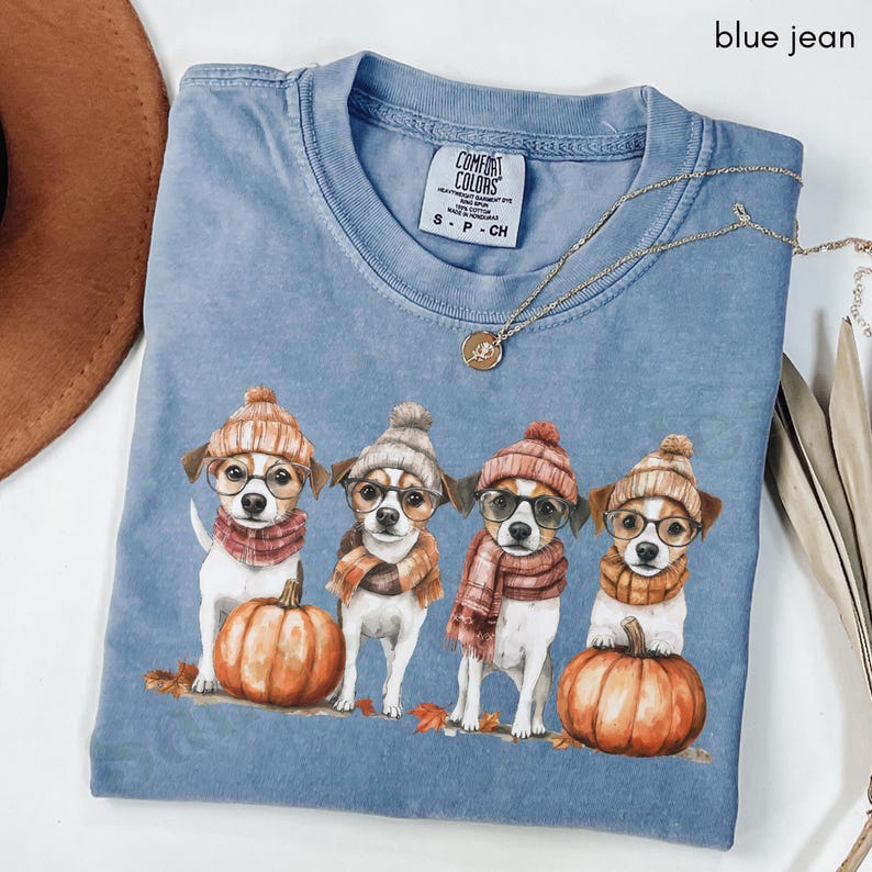 K&ouml;nnte beinhalten: Blaues Comfort Colors T-Shirt mit einem Aufdruck von vier Hunden, die H&uuml;te, Schals und Brillen tragen und neben K&uuml;rbissen stehen. Das Shirt hat einen Rundhalsausschnitt und kurze &Auml;rmel. Eine goldene Halskette ist &uuml;ber das Shirt drapiert.