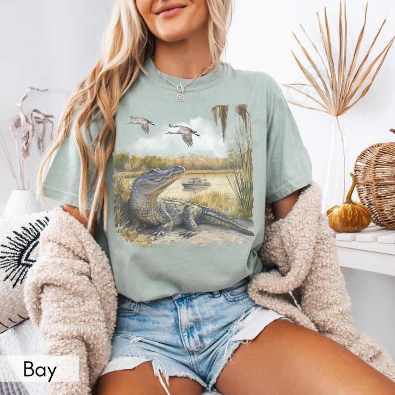 May include: A light green t-shirt featuring a detailed illustration of an alligator in a swamp setting. The artwork includes flying birds, a boat, and a natural landscape. The word "Bay" is printed at the bottom of the image.