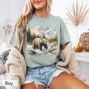 Puede incluir: Una camiseta verde claro con un gr&aacute;fico de un oso marr&oacute;n caminando por un paisaje monta&ntilde;oso nevado. El oso camina por un arroyo con rocas a cada lado. Las monta&ntilde;as est&aacute;n en el fondo con un cielo azul y nubes blancas.