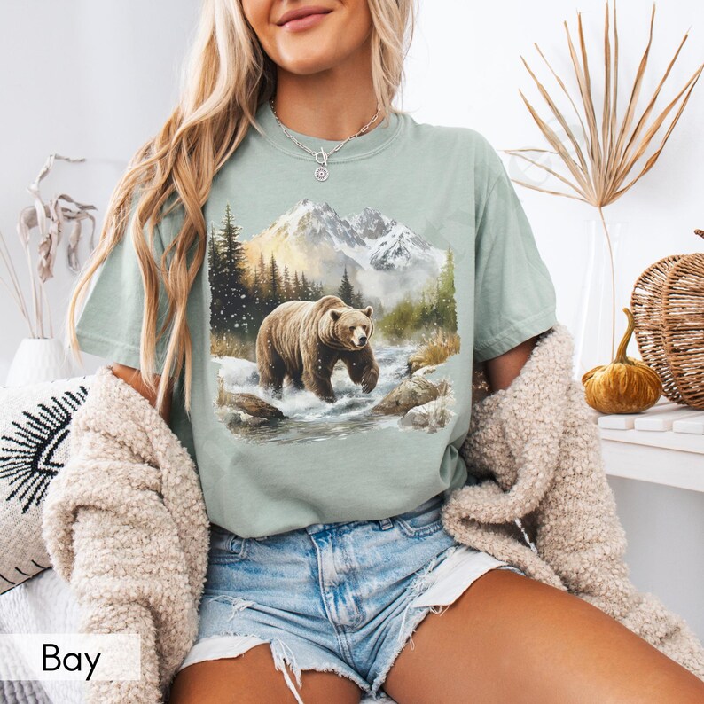 Puede incluir: Una camiseta verde claro con un gr&aacute;fico de un oso marr&oacute;n caminando por un paisaje monta&ntilde;oso nevado. El oso camina por un arroyo con rocas a cada lado. Las monta&ntilde;as est&aacute;n en el fondo con un cielo azul y nubes blancas.