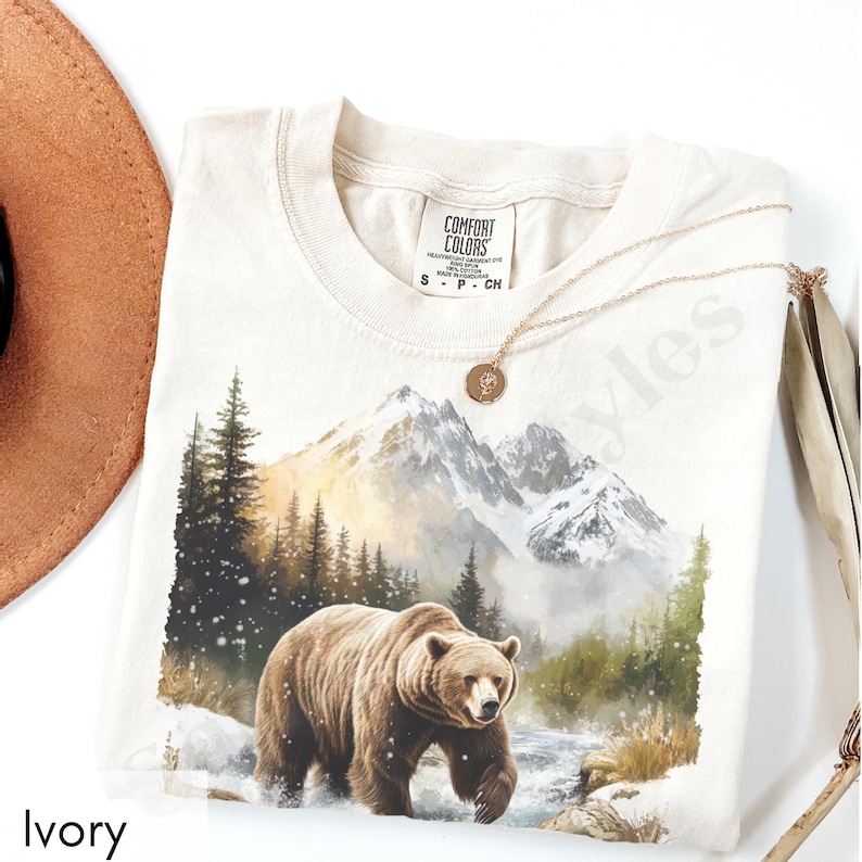 Puede incluir: Una camiseta beige con un estampado gr&aacute;fico de un oso marr&oacute;n caminando por un paisaje monta&ntilde;oso nevado. La etiqueta dice "COMFORT COLORS". La camiseta est&aacute; etiquetada como "Ivory".