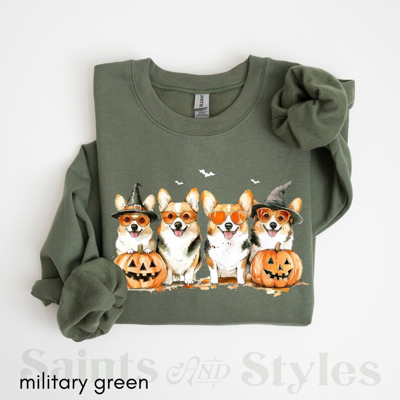 May include: A green sweatshirt with a Halloween-themed design featuring four corgis wearing witch hats and sunglasses, holding pumpkins. The corgis are surrounded by bats and fall leaves.