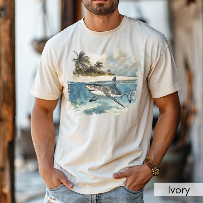 May include: A white t-shirt with a graphic of a great white shark swimming in the ocean near a tropical island. The shirt is ivory in color.