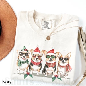 May include: Ivory t-shirt featuring a graphic of four corgis wearing Santa hats and sunglasses. The corgis are wearing scarves and are standing in a row. The text "COMFORT COLORS" is on the tag.