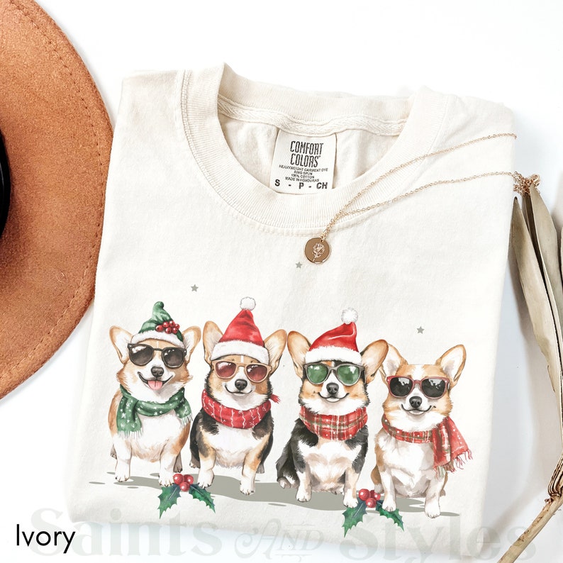May include: Ivory t-shirt featuring a graphic of four corgis wearing Santa hats and sunglasses. The corgis are wearing scarves and are standing in a row. The text "COMFORT COLORS" is on the tag.