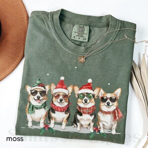 May include: A green t-shirt with a Christmas-themed design featuring four corgis wearing Santa hats and sunglasses. The corgis are wearing scarves and are standing in a row. The text "COMFORT COLORS" is visible on the tag.