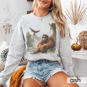 Op de afbeelding: Een grijs sweatshirt met een grafische print van een orang-oetan die in een jungle-omgeving zit met een blauw-rode papegaai die erboven vliegt. De tekst "ash" is gedrukt in de rechterbenedenhoek van de afbeelding.