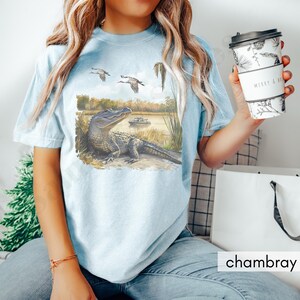 May include: Light blue t-shirt featuring a watercolor illustration of an alligator resting near a lake, with flying birds and a boat in the background. The shirt has the word "chambray" printed on it.