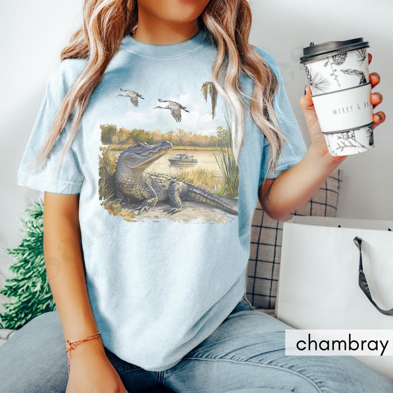 May include: Light blue t-shirt featuring a watercolor illustration of an alligator resting near a lake, with flying birds and a boat in the background. The shirt has the word "chambray" printed on it.