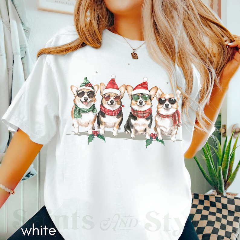 May include: White t-shirt featuring a design of four corgis wearing Santa hats and sunglasses. The corgis are wearing scarves and are standing in a line.