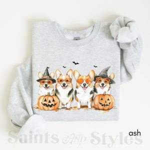 May include: A gray sweatshirt with a design of four corgis wearing witch hats and sunglasses, holding jack-o'-lanterns. The corgis are in a row, facing forward. The sweatshirt is on a white background.