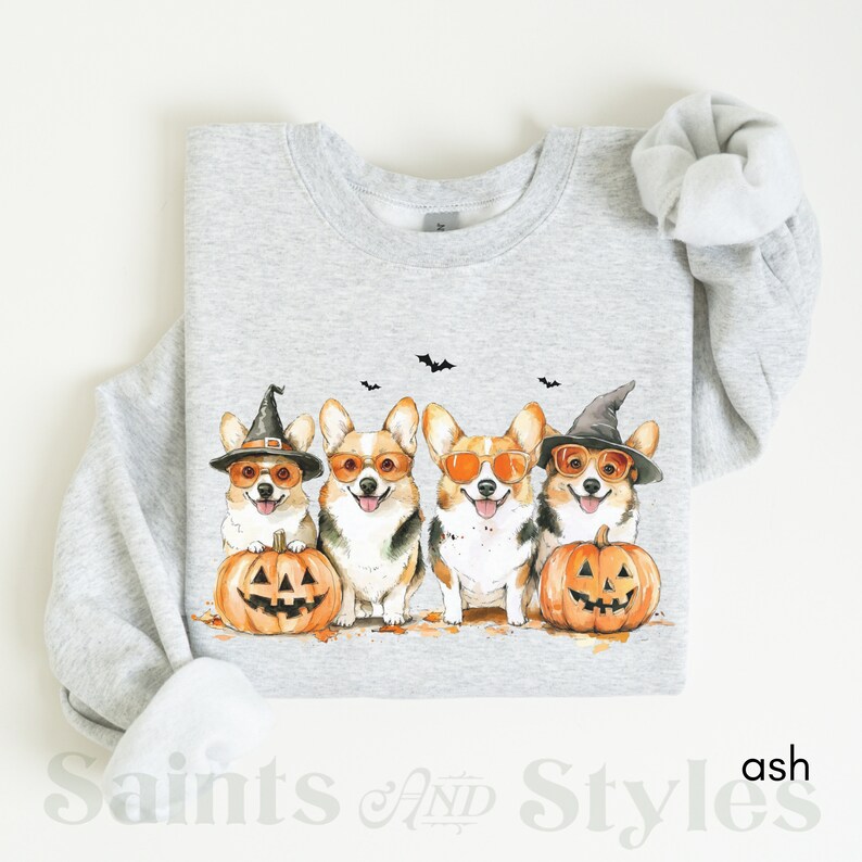 May include: A gray sweatshirt with a design of four corgis wearing witch hats and sunglasses, holding jack-o'-lanterns. The corgis are in a row, facing forward. The sweatshirt is on a white background.