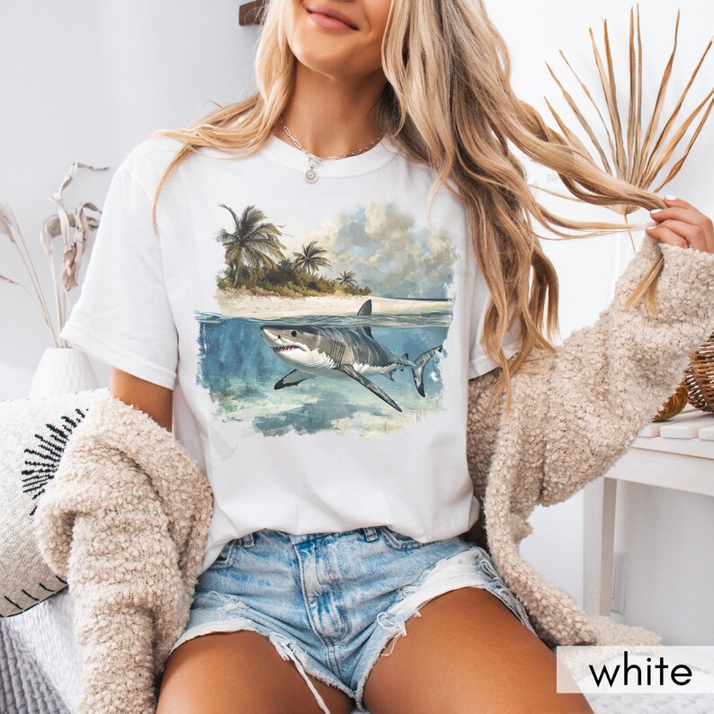 May include: White t-shirt with a graphic of a shark swimming in the ocean with palm trees on a tropical island in the background.