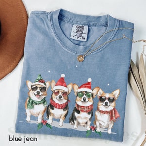May include: Blue jean t-shirt featuring a Christmas design with four corgis wearing Santa hats and sunglasses. The corgis are wearing scarves and are standing in a row on a snowy background. The shirt has a tag that says "Comfort Colors" and "S-P-CH".