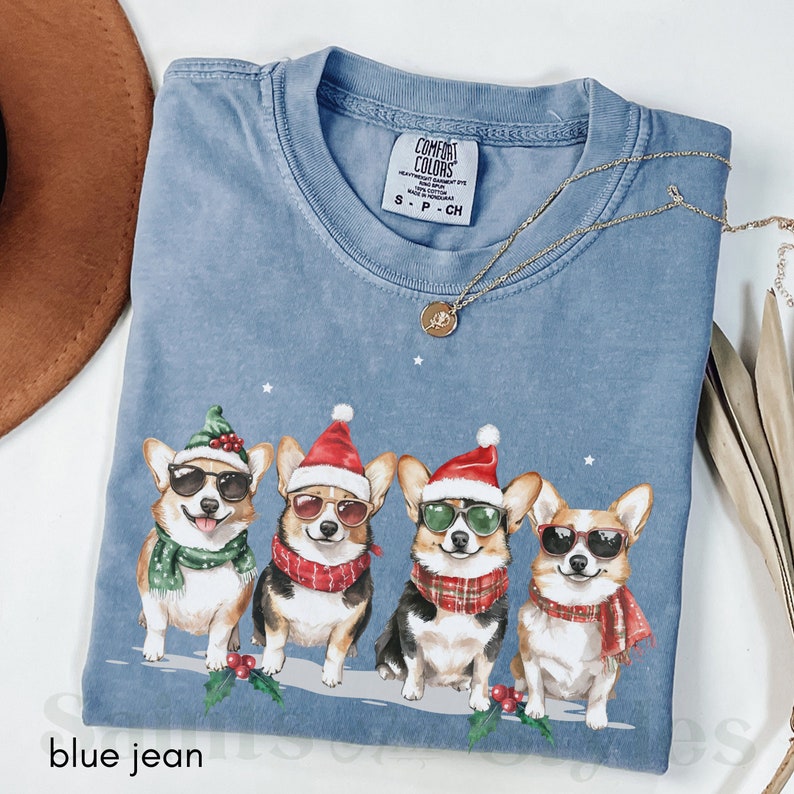 May include: Blue jean t-shirt featuring a Christmas design with four corgis wearing Santa hats and sunglasses. The corgis are wearing scarves and are standing in a row on a snowy background. The shirt has a tag that says "Comfort Colors" and "S-P-CH".