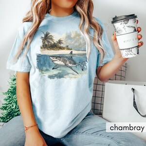 May include: A light blue t-shirt with a graphic of a great white shark swimming in the ocean. The shark is white with a gray shadow. The ocean is a light blue with a white foamy wave. There are palm trees on a small island in the background.