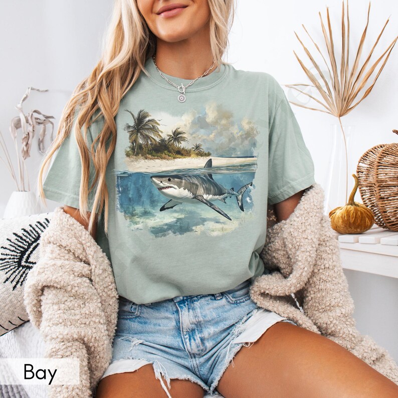 May include: A light blue t-shirt with a graphic of a white shark swimming in the ocean. The shark is swimming towards the viewer. The graphic also includes a small island with palm trees in the background. The shirt has a crew neck and short sleeves.