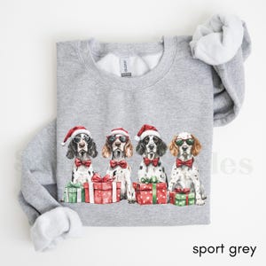 May include: A gray sweatshirt with a Christmas-themed design featuring four dogs wearing Santa hats and sunglasses. The dogs are holding presents wrapped in red and green paper.