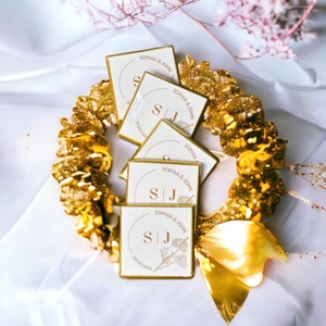 Op de afbeelding: Een gouden krans omringt verschillende witte vierkante gunsten met gouden rand. Elke gunst is bedrukt met de namen "SOPHIA & JOHN", initialen "SJ" en de datum "20/05/2025". De gunsten zijn gerangschikt op een wit oppervlak.
