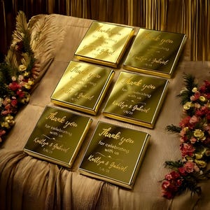 Könnte beinhalten: Sechs goldfarbene, rechteckige Schachteln mit dem eingravierten Text "Thank you for celebrating with us Kaitlyn & Gabriel 7/8/25" auf der Oberseite. Die Schachteln sind auf einer beigen Oberfläche angeordnet, mit Blumenarrangements auf beiden Seiten.
