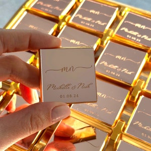 K&ouml;nnte beinhalten: Eine Schachtel mit kleinen, goldverpackten Pralinen mit rosafarbener Innenseite. Die Pralinen sind mit den Initialen "MN" und den Namen "Michelle & Nish" sowie dem Datum "01.08.24" personalisiert.