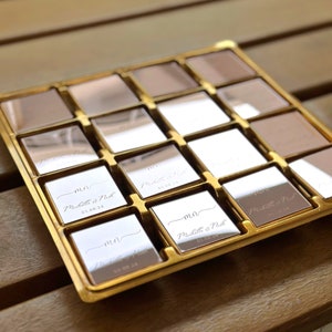 May include: A gold-coloured tray containing sixteen square, mirrored tiles. Each tile is engraved with the initials "MN", the names "Michelle & Nick", and the date "01.08.24". The tiles have a reflective surface.