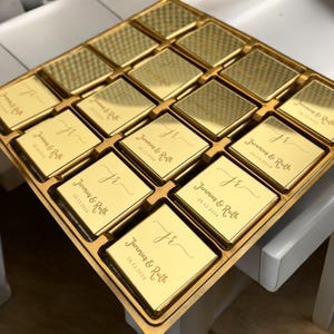 May include: A gold-coloured box filled with square chocolates. Each chocolate is engraved with the initials "JR", the names "Jeremias & Ruth", and the date "26.12.2024". The chocolates are arranged in a grid pattern.