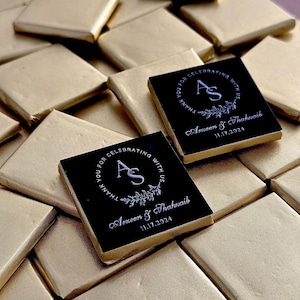 May include: Square chocolates wrapped in gold foil. Two chocolates with black tops feature white text: "THANK YOU FOR CELEBRATING WITH US" around a monogram "AS", names, and the date 11.17.2024. The chocolates are for a wedding.
