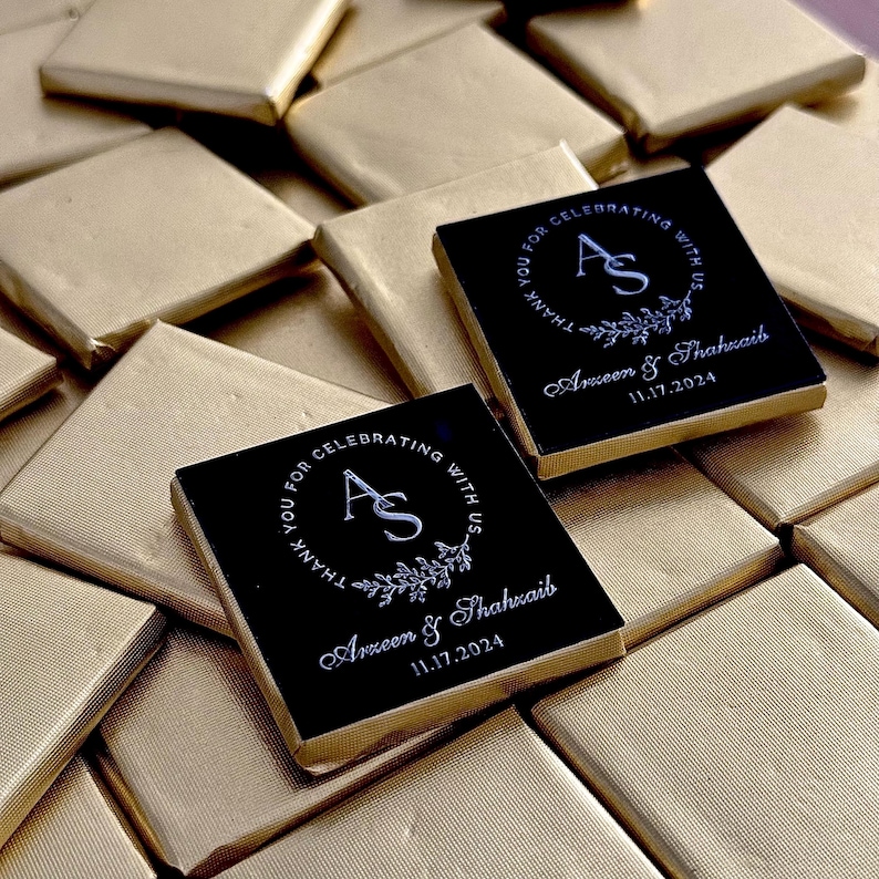 May include: Square chocolates wrapped in gold foil. Two chocolates with black tops feature white text: "THANK YOU FOR CELEBRATING WITH US" around a monogram "AS", names, and the date 11.17.2024. The chocolates are for a wedding.