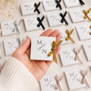 May include: A collection of square white favour boxes featuring the printed letters M and N separated by a small botanical illustration. Each box is tied with a small satin ribbon in various colours including gold, black, silver, and beige.
