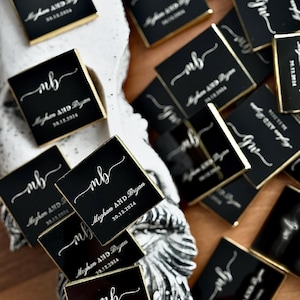 May include: Black square favors with gold edging, each displaying the initials "mb" in white script. The text "Meghan AND Bryan" and the date "30.12.2024" are printed on the favors. The favors are arranged on a textured surface.