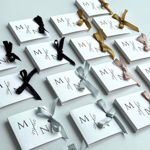 May include: White square cards printed with the letters "M" and "N" in brown, and a botanical design. Each card is tied with a ribbon in various colours, including black, gold, and silver. The cards are arranged on a white surface.