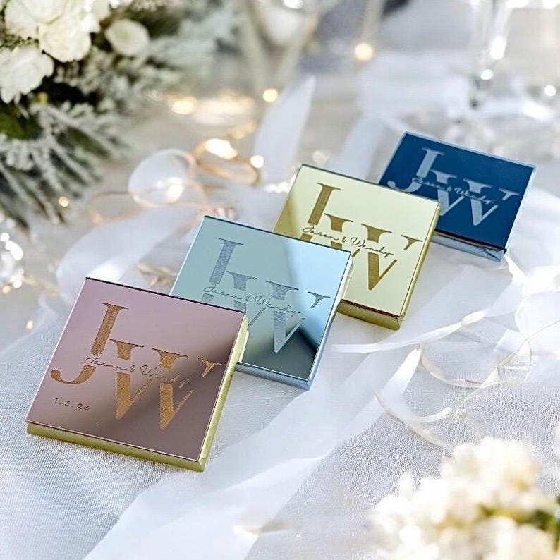 May include: Four square, mirrored boxes with gold lettering. The boxes are in different colors: rose gold, silver, gold, and blue. The lettering on the boxes reads 'J&W' and 'Jean & Wendy' with the date '1.5.24' on the rose gold box.