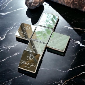 May include: Silver mirrored boxes arranged in a cross shape, each engraved with a wedding ring design, names, and a date. The boxes reflect the surrounding environment. The background is a black marble surface.